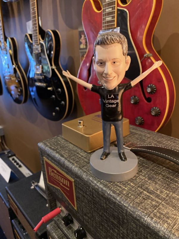 A smiling Clif Jones bobblehead with outstretched arms among guitars and amps